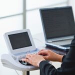 A person is sitting at a desk, typing on a stenotype machine. Next to the device is a laptop.