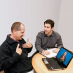 Two men are sitting at a table, with a binder and laptop between them. The man on the left is signing in ASL and looking at the laptop. On the laptop screen is an ASL interpreter.