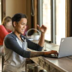 A woman in a henley tee, with short brown hair, is sitting in a café, smiling and signing in ASL at her laptop screen. In the background of the image is a purple ribbon.