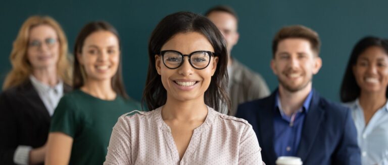 Focused in the middle of the image is a woman with black hair, wearing glasses and a v-neck blouse, is standing and smiling. Behind her, out of focus, is a group of people, wearing business casual clothing, all standing next to each other and smiling.