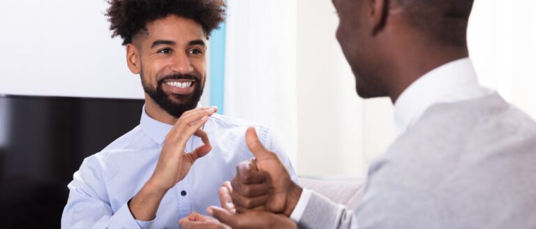 A Black man with curly black hair, wearing a purple button-down shirt, is in an office, signing in ASL and smiling at a man in front of him. The man in front of him is wearing a grey sweater and white button-down shirt, smiling and signing in ASL.