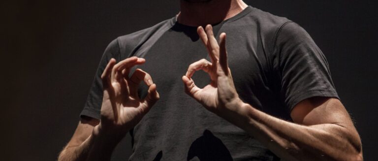 A close-up image of a man wearing a black t-shirt signing in ASL.