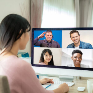 A woman is sitting at a desk, looking at her computer monitor. On the computer screen is a video meeting of four people. One of the people in the meeting is an interpreter. The interpreter is signing and smiling.