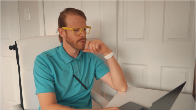 A white man with brown hair in a ponytail and brown beard, wearing a blue polo top and yellow glasses, sitting at a desk, looking at his laptop screen.