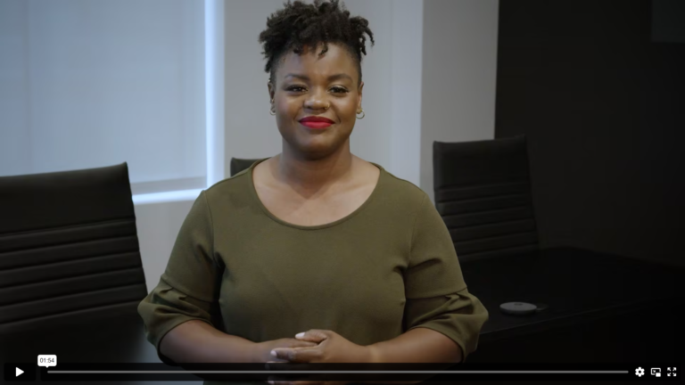 A Black woman with short black hair, wearing an olive sweater, is smiling. Behind her is an office table with multiple black office chairs.