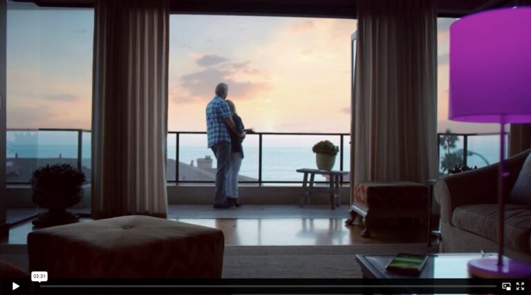 Thumbnail of a video shows a couple in an embrace, looking at the sunset on a balcony outside of a living room.