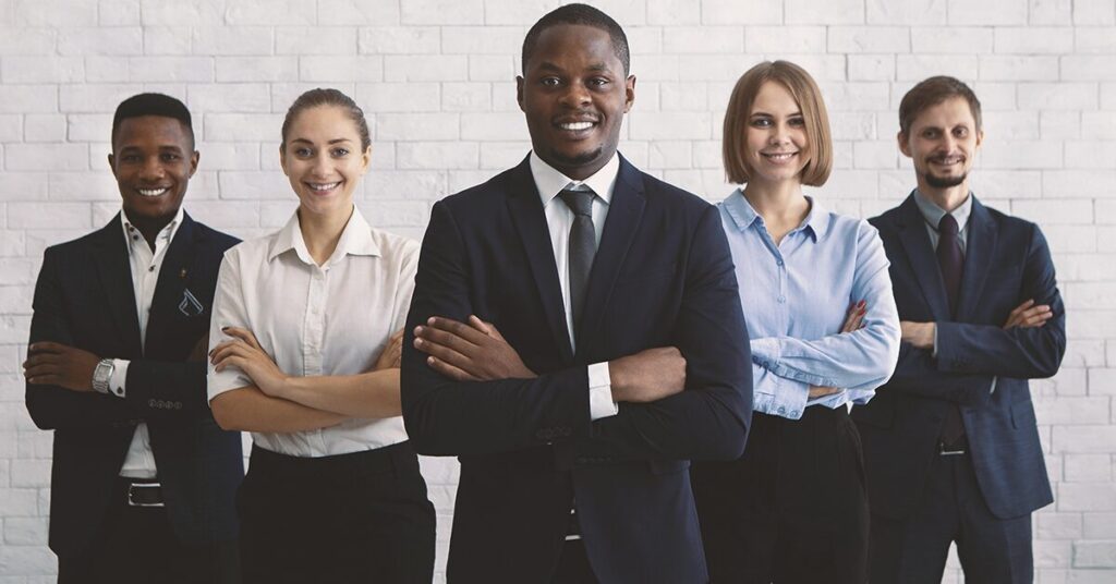 The image shows five people in business casual clothing, with their arms folded, smiling and looking at the camera. Behind them is a white brick wall.