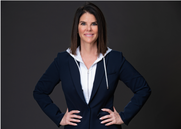 A woman, ZP CEO Sherri Turpin, with short brown hair is smiling and standing in front of a black background, with her hands on her hips, wearing a grey zip-up jacket with a blazer.