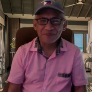 Hector Brual A man is sitting on a chair, wearing a grey hat, pink short sleeve button-down and black glasses, smiling.