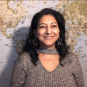 Kamini Dalal A woman with black hair, wearing a grey sweater and dangling earrings, is smiling. Behind her is a world map.