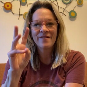 Peggy Ann Wegner A white woman with blonde hair, wearing a red graphic tee and glasses, is signing and smiling.