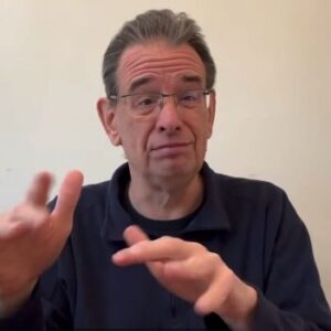 pat-costello-screenshot A white man with grey hair and glasses, wearing a navy collared long sleeve top, is signing in ASL.